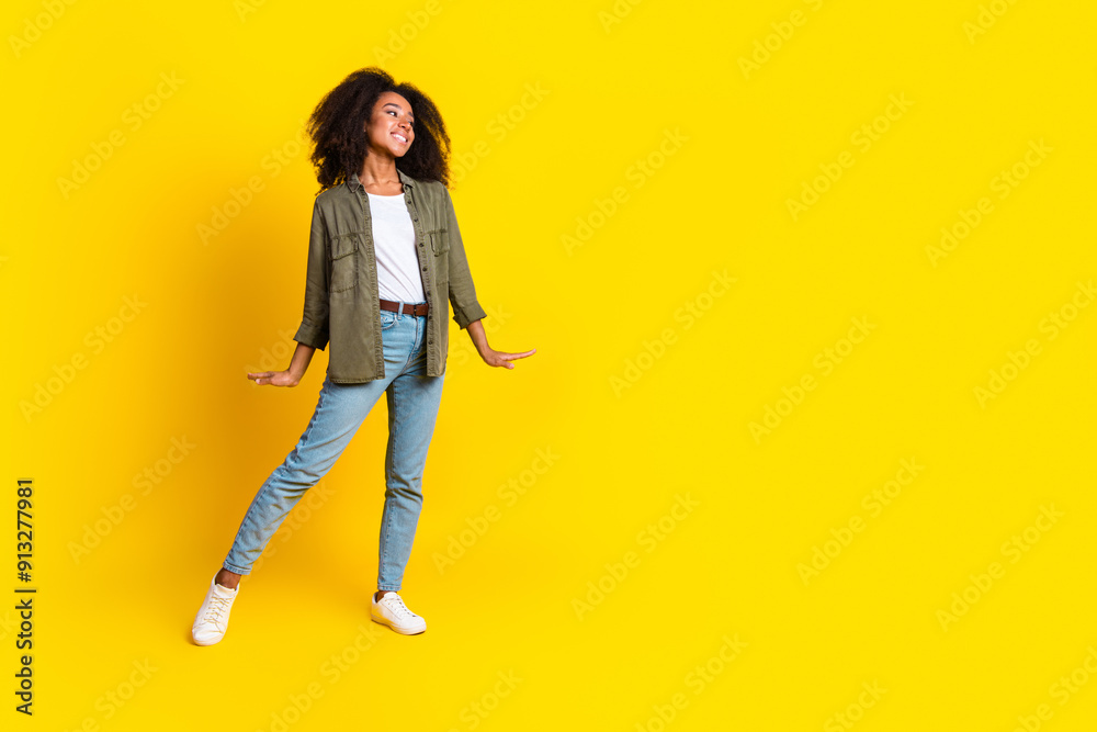 Full size photo of lovely girl dressed khaki jacket jeans pants dancing look at logo empty space isolated on yellow color background Full size photo of lovely girl dressed khaki jacket jeans pants dancing look at logo empty space isolated on yellow color background