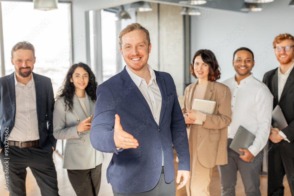 A group of diverse business professionals are standing in a modern office, with one man extending his hand for a handshake. The others are smiling and looking at the camera. A group of diverse business professionals are standing in a modern office, with one man extending his hand for a handshake. The others are smiling and looking at the camera.