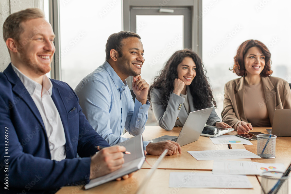 A group of four business professionals are gathered around a table in a modern office setting. They are engaged in a meeting, listening attentively and taking notes. The atmosphere is positive