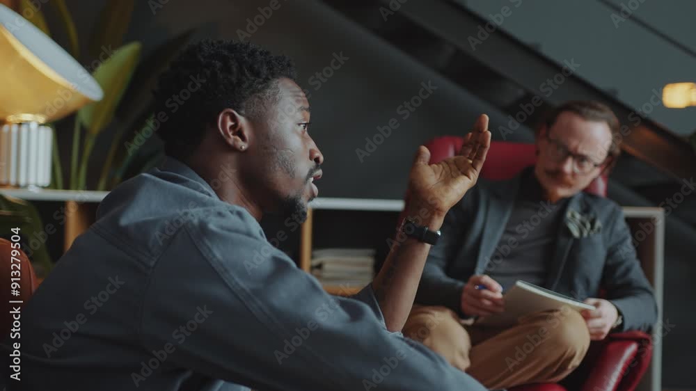 Young Black man explaining his feelings to Caucasian male therapist listening attentively, writing notes and giving advice during individual counseling session in cozy office. Rack focus