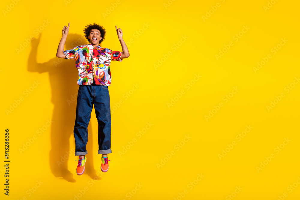 Full size photo of nice funky young man jump direct fingers up empty space wear shirt isolated on bright yellow color background Full size photo of nice funky young man jump direct fingers up empty space wear shirt isolated on bright yellow color background