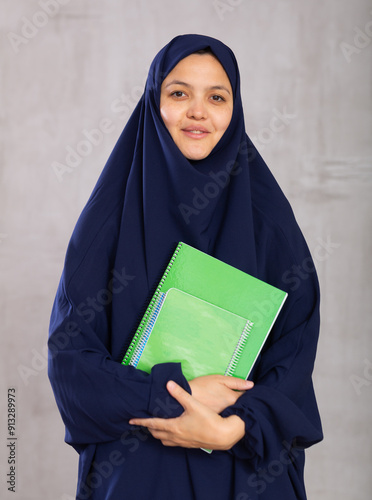 Portrait of young smiling female student in hijab posing with notebooks in studio