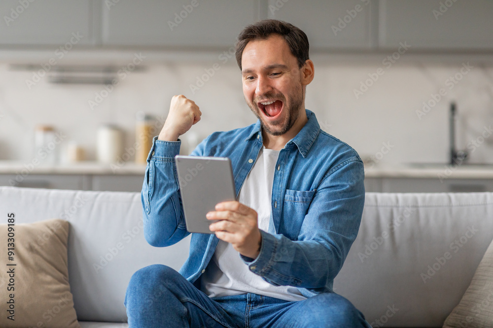 A man sits on a white couch in a living room and celebrates with a raised fist while looking at a tablet in his hands. He is wearing a white t-shirt and a blue denim shirt. A man sits on a white couch in a living room and celebrates with a raised fist while looking at a tablet in his hands. He is wearing a white t-shirt and a blue denim shirt.
