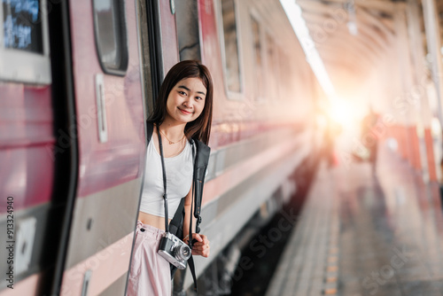 A young woman with a camera leans out of a train at a station platform, smiling as the sun sets in the background.