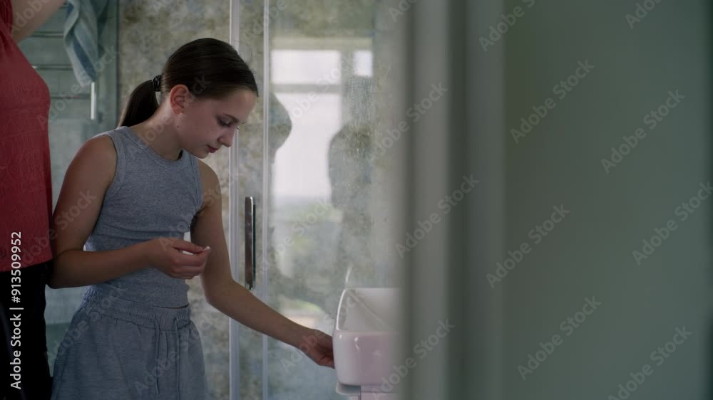 Adult woman looks at her reflection in the mirror and dries wet hair after shower. Young girl stands near mother in bathroom and makes skincare procedures. Concept of morning bath routine. Close up. Adult woman looks at her reflection in the mirror and dries wet hair after shower. Young girl stands near mother in bathroom and makes skincare procedures. Concept of morning bath routine. Close up.