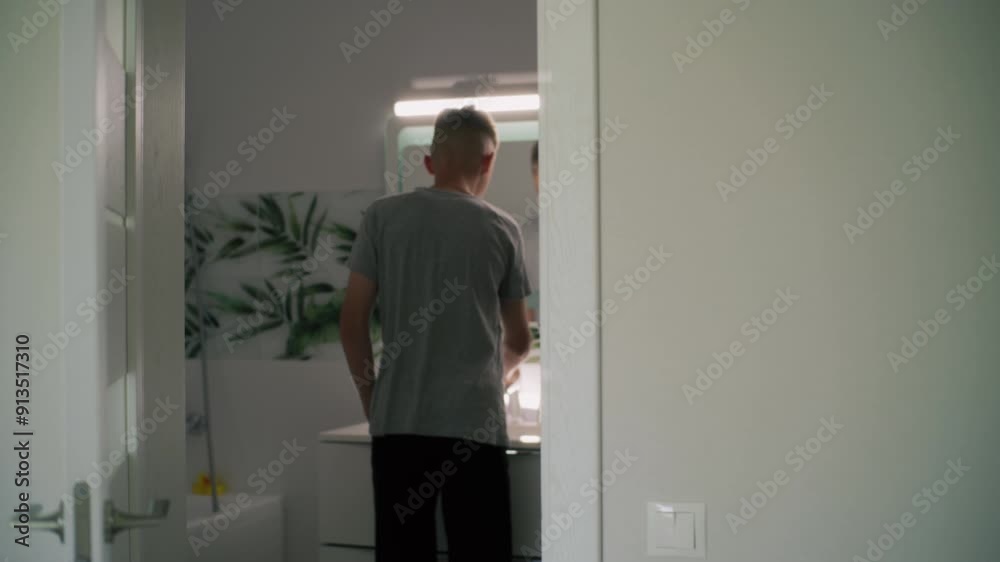 Caucasian boy comes into bathroom, washes face with cold water and looks at his reflection in the mirror. Teenager stands in front of basin and makes morning bathroom routine. Back view through door. Caucasian boy comes into bathroom, washes face with cold water and looks at his reflection in the mirror. Teenager stands in front of basin and makes morning bathroom routine. Back view through door.