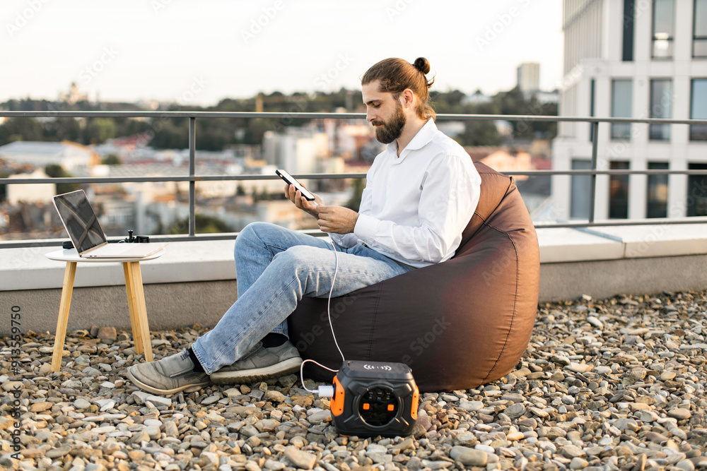 Businessman sitting on beanbag chair and plugs phone charging cable into portable power station on rooftop of house. Laptop on table nearby, urban background. Concept of remote work, technology. Businessman sitting on beanbag chair and plugs phone charging cable into portable power station on rooftop of house. Laptop on table nearby, urban background. Concept of remote work, technology.