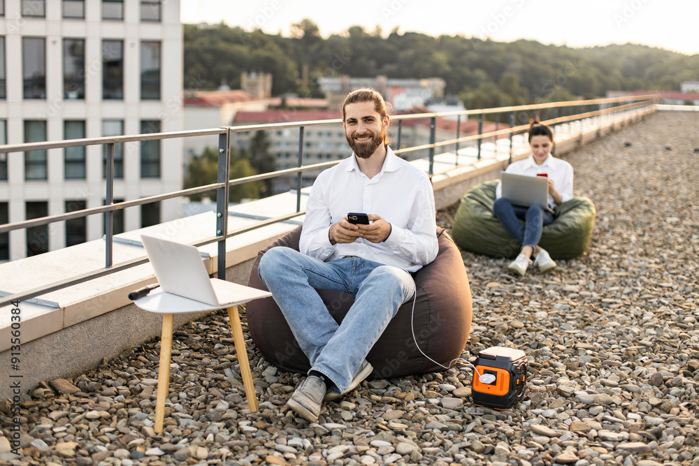 Businessman sitting on beanbag chair and plugs phone charging cable into portable power station on roof of house. Another person uses laptop in background Businessman sitting on beanbag chair and plugs phone charging cable into portable power station on roof of house. Another person uses laptop in background