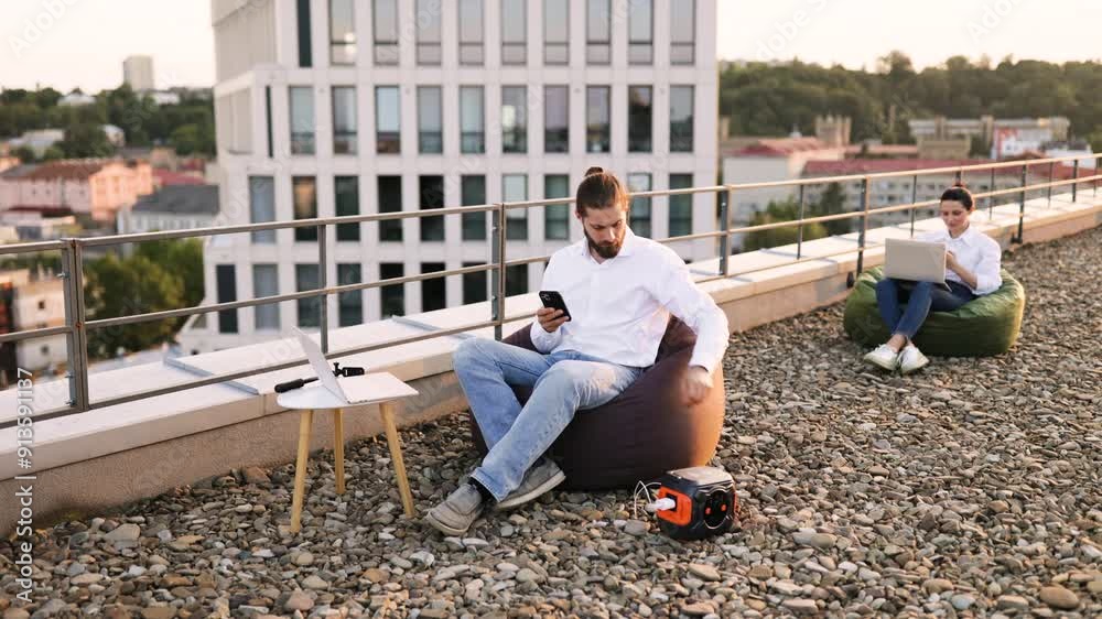 Businessman sitting on beanbag chair and plugs phone charging cable into portable power station on roof of house. Another person working on laptop in background. Businessman sitting on beanbag chair and plugs phone charging cable into portable power station on roof of house. Another person working on laptop in background.