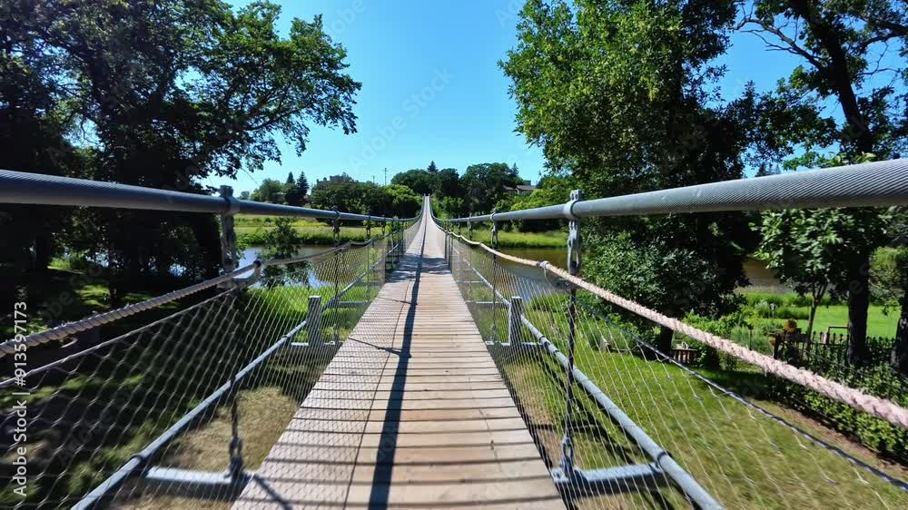 POV shot walking across a suspension bridge on a sunny day. Lush green trees line the riverbanks. POV shot walking across a suspension bridge on a sunny day. Lush green trees line the riverbanks.
