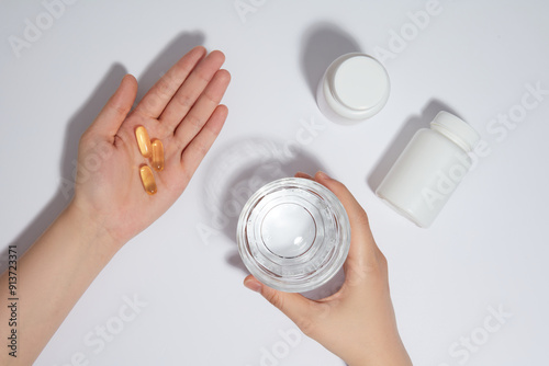 Photo scene of the preparation for medicine drinking on white background from top angle. Female's hand with some yellow pills in palm, the other one holding a water glass, next to some unlabeled jars