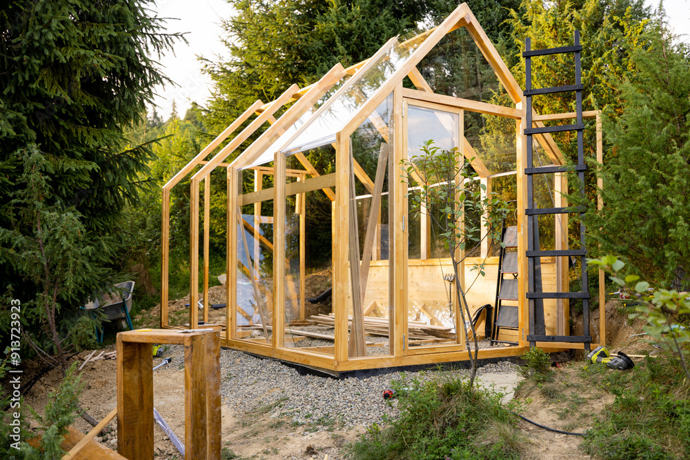 A wooden greenhouse under construction in a lush backyard surrounded by tall trees. The scene captures the early stages of a DIY project in an eco-friendly environment, highlighting sustainable living