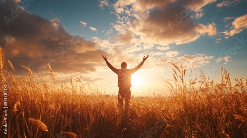 A man stands in a golden field, arms raised high in prayer, basking in the warm glow of a stunning sunset with clouds painted in vibrant colors