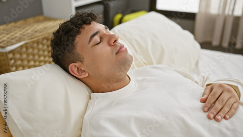 A young man with curly hair sleeps peacefully in a bright bedroom, lying on white bedding indicating relaxation and comfort.