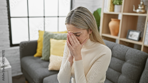 Young blonde woman looks distressed, covering face with hand, in a cozy living room setting.