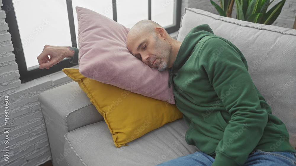 A bald man with a beard, wearing a green sweatshirt, naps on colorful cushions on a couch indoors, suggesting relaxation at home. A bald man with a beard, wearing a green sweatshirt, naps on colorful cushions on a couch indoors, suggesting relaxation at home.