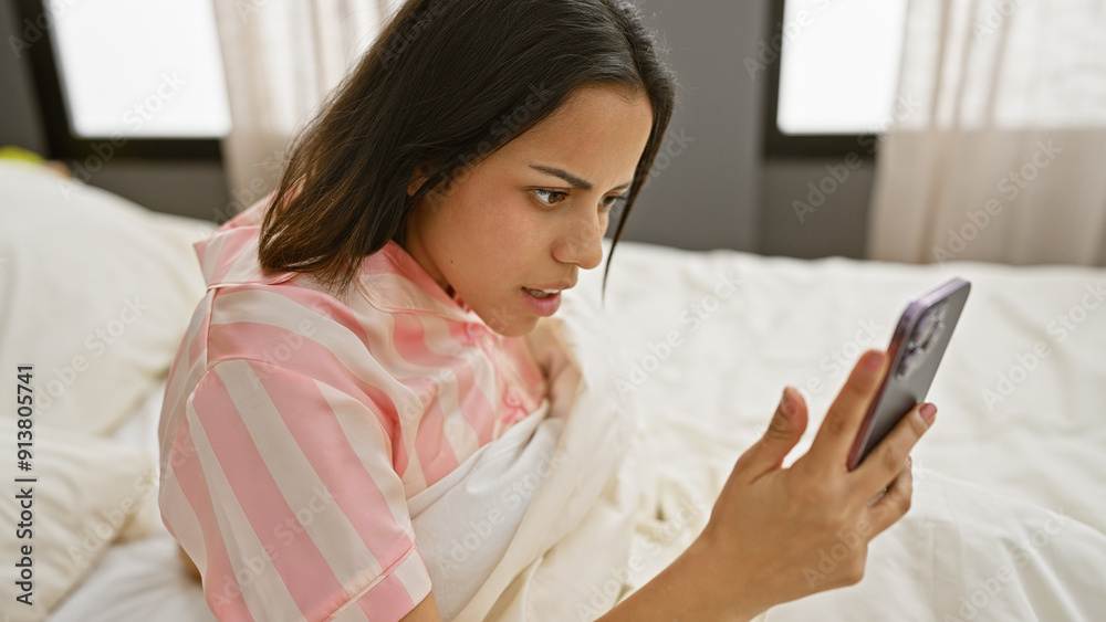 A young hispanic woman in pajamas focusing on a smartphone in a bright cozy bedroom A young hispanic woman in pajamas focusing on a smartphone in a bright cozy bedroom