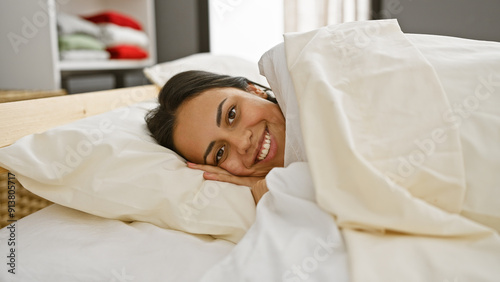 A smiling young hispanic woman comfortably relaxes in a cozy bedroom, exuding an air of warmth and tranquility.