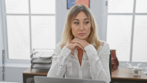 A contemplative caucasian woman sits in an office, with a laptop, papers, drone on desk and a bright window behind.