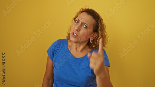 Slika na platnu Mature hispanic woman with curly hair in a blue shirt pointing and giving a stern expression, isolated against a yellow background