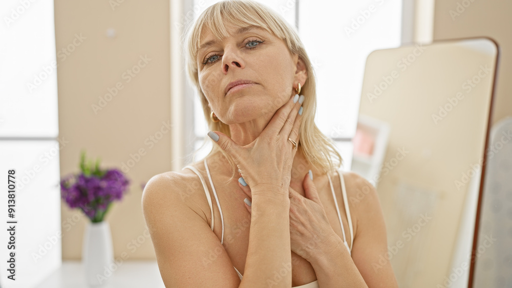 A contemplative blonde woman touches her neck in a bright living room with a mirror and flowers in the background. A contemplative blonde woman touches her neck in a bright living room with a mirror and flowers in the background.