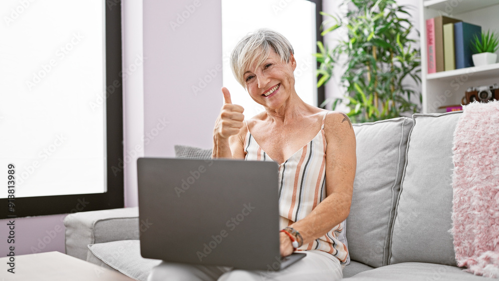 A smiling mature woman with grey hair gives a thumbs-up while using a laptop on a sofa indoors. A smiling mature woman with grey hair gives a thumbs-up while using a laptop on a sofa indoors.