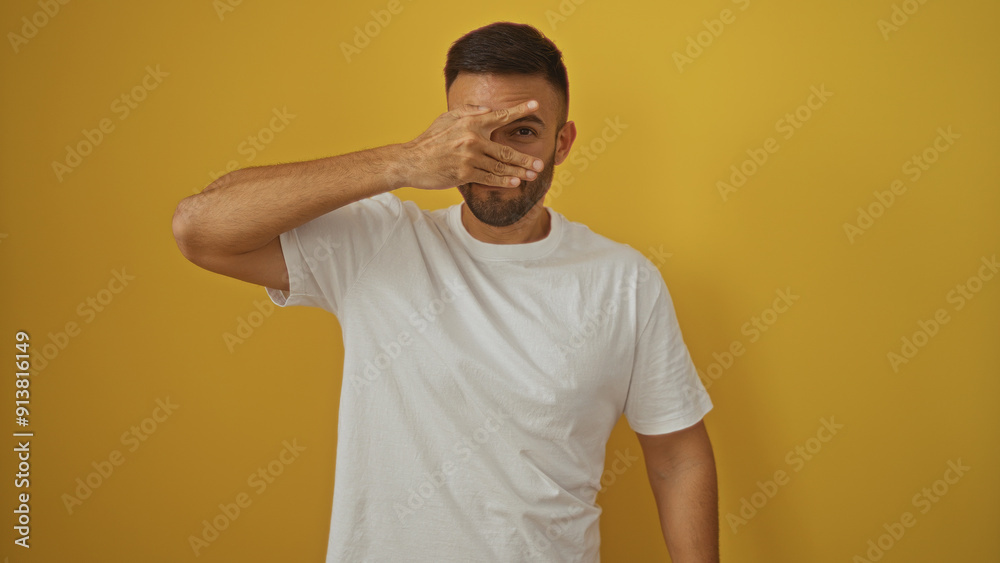 A handsome young hispanic man with a beard in a white shirt poses over an isolated yellow background, covering one eye with his hand for a playful portrait.