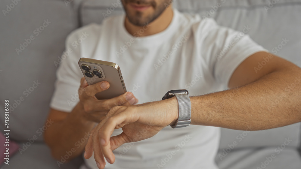 A handsome hispanic man with a beard interacts with his smartphone and smartwatch while lounging in a modern apartment's living room. A handsome hispanic man with a beard interacts with his smartphone and smartwatch while lounging in a modern apartment's living room.