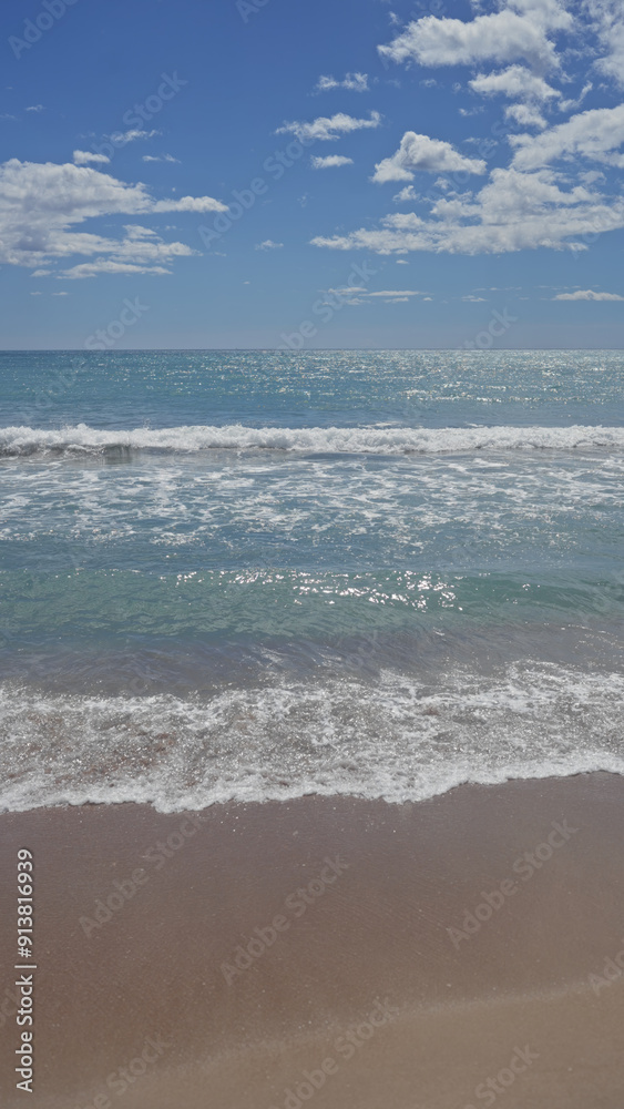 A serene beach with sparkling turquoise waters, white waves, sandy shore under a sunny sky with scattered clouds.