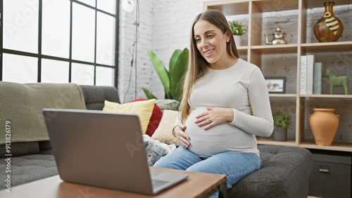 Quadro em tela A smiling pregnant hispanic woman sits comfortably at home with a laptop, exuding maternal warmth in a cozy living room