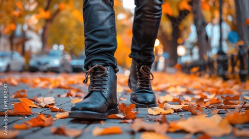 A person wearing black leather boots and jeans walks along a sidewalk covered in fallen autumn leaves. The leaves are a bright orange, and the sunlight creates a warm glow on the scene.