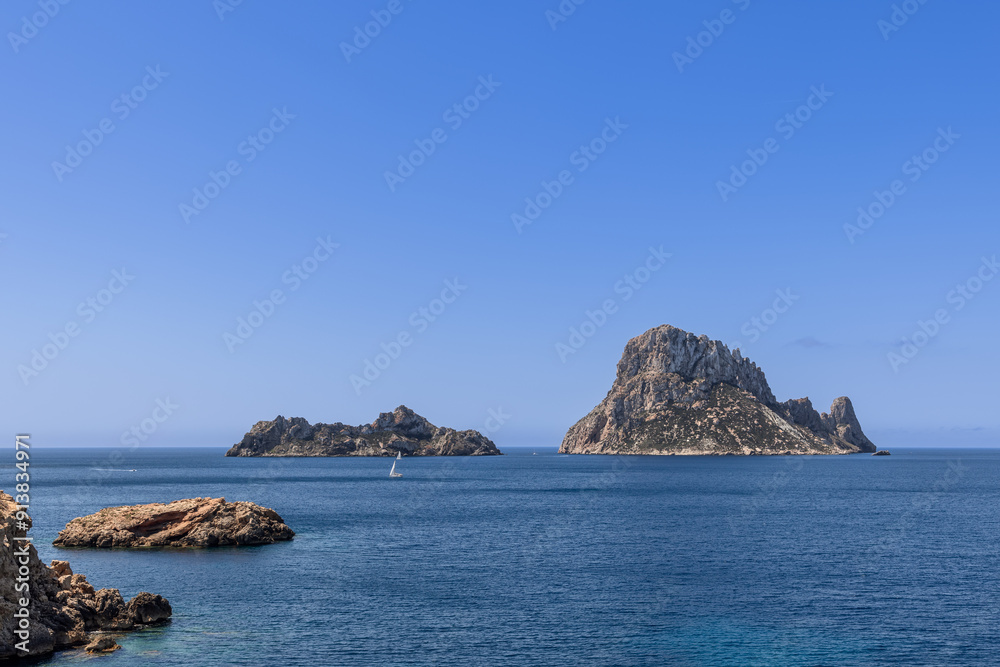 Scenic panorama of Es Vedra and Es Vedranell islets in Ibiza, Spain. The azure waters of the Mediterranean stretch out, with a solitary sailboat navigating the serene sea under a bright blue sky Scenic panorama of Es Vedra and Es Vedranell islets in Ibiza, Spain. The azure waters of the Mediterranean stretch out, with a solitary sailboat navigating the serene sea under a bright blue sky