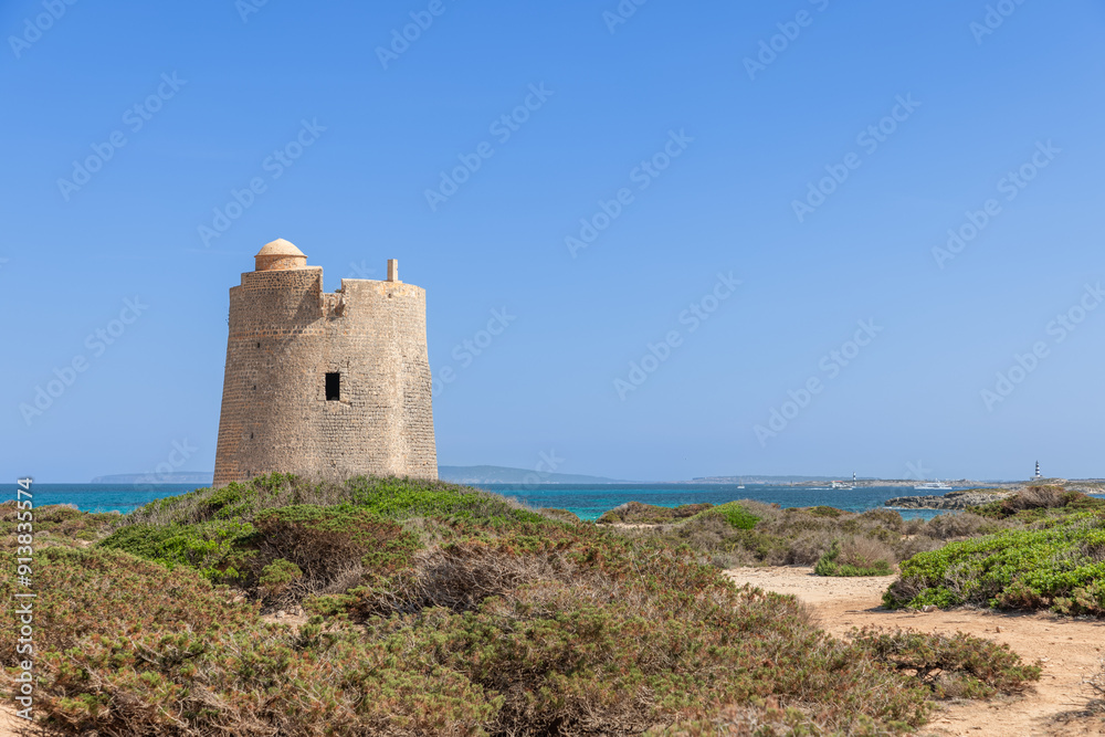 The ancient coastal tower Torre de ses Portes on Ibiza stands proudly against the turquoise sea, surrounded by lush coastal vegetation and a clear blue sky The ancient coastal tower Torre de ses Portes on Ibiza stands proudly against the turquoise sea, surrounded by lush coastal vegetation and a clear blue sky