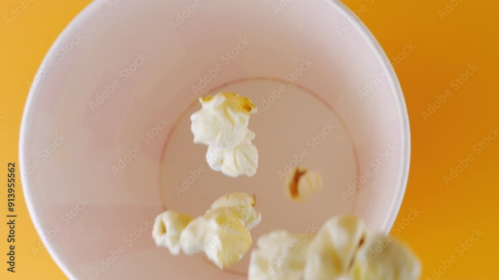Popcorn grains falling into red and white striped bucket isolated on peach background. Movie snack food, fast food cafe sweet snack. Top view. Slow motion. Popcorn grains falling into red and white striped bucket isolated on peach background. Movie snack food, fast food cafe sweet snack. Top view. Slow motion.