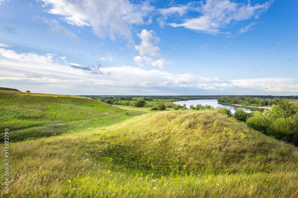 Rolling green hills gently slope toward a calm river, under a bright blue sky scattered with fluffy white clouds, creating a peaceful and natural oasis. Rolling green hills gently slope toward a calm river, under a bright blue sky scattered with fluffy white clouds, creating a peaceful and natural oasis.