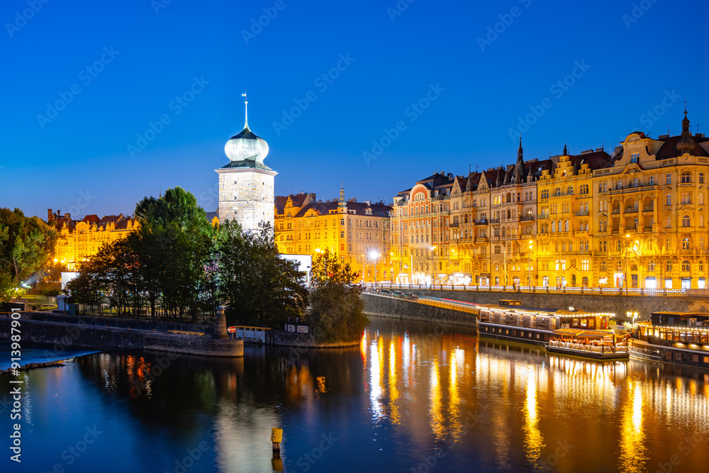 Fototapeta premium The Sitkov Water Tower stands illuminated on the Masaryk Embankment in Prague, Czechia, as dusk descends. The tower is reflected in the tranquil waters of the Vltava River.