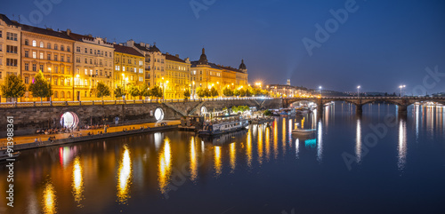 A view of the Rasin Embankment and Palacky Bridge in Prague, Czechia at dusk. The bridge is lit up, and the city lights reflect in the water. There are boats on the river, and the sky is a deep blue. photo