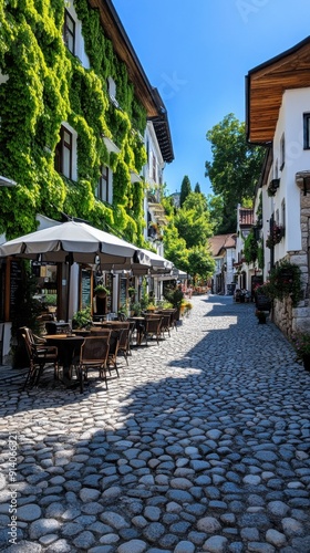 Charming cobblestone street with lush greenery and inviting cafe terraces, perfect for leisurely strolls and outdoor dining.