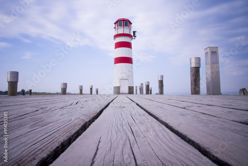A red and white lighthouse stands at the end of a wooden pier overlooking the ocean under a clear sky.