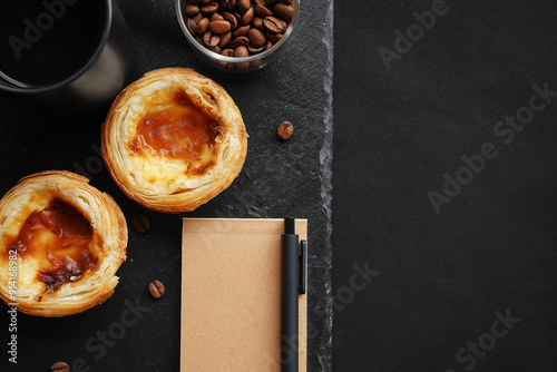 Egg tarts next to a black cup of coffee, scattered coffee beans and a notepad on a dark background photo