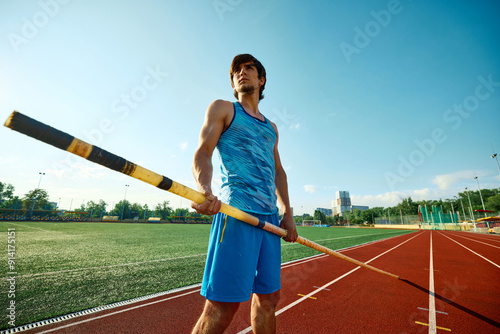 Tableau sur toile Bottom view portrait of focused and determined pole vaulter, athlete in blue uniform standing with pole at open air stadium, showing readiness to train