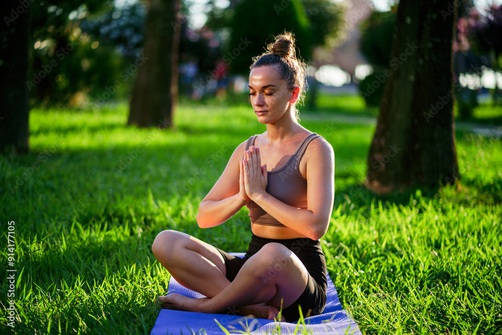 Concentrated young woman with crossed legs, praying hands and closed eyes in lotus position meditating on yoga mat in park at sunset, breathing exercise Concentrated young woman with crossed legs, praying hands and closed eyes in lotus position meditating on yoga mat in park at sunset, breathing exercise