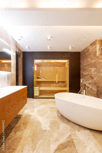 Modern bathroom with marble walls and floor. Bathtub in the middle of the room. In the background you can see a sauna with glass door.
