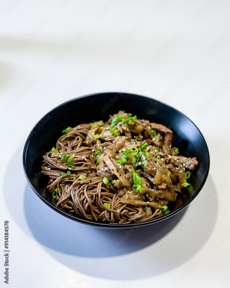 Buckwheat soba noodles with pork and sweet and sour sauce in a black plate on a white table Buckwheat soba noodles with pork and sweet and sour sauce in a black plate on a white table
