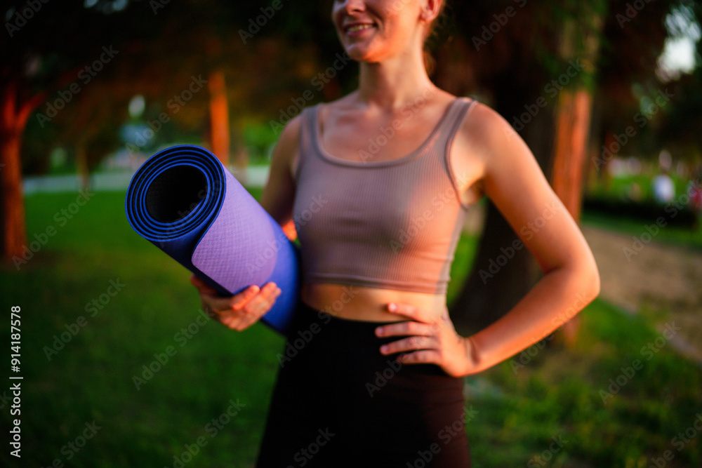 Slim young woman holding rolled fitness mat in park at sunset, wellbeing and healthy sportive lifestyle Slim young woman holding rolled fitness mat in park at sunset, wellbeing and healthy sportive lifestyle