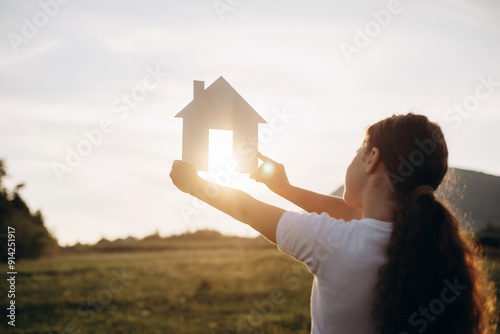 Close-up of preteen girl holding paper house model on background beautiful warm sunset and amazing mountains. Home in rays of sun in summer nature. Dream own house. Housing and mortgage concept