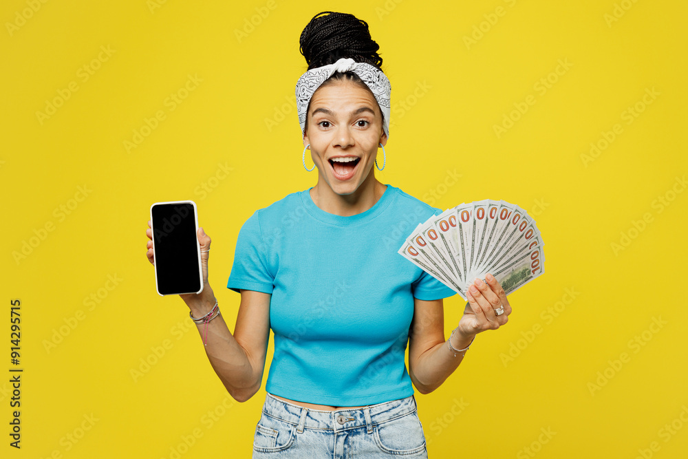 Young Latin woman wears blue top t-shirt headband casual clothes use blank screen mobile cell phone hold fan of cash money in dollar banknotes isolated on plain yellow background. Lifestyle concept. Young Latin woman wears blue top t-shirt headband casual clothes use blank screen mobile cell phone hold fan of cash money in dollar banknotes isolated on plain yellow background. Lifestyle concept.