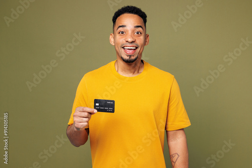 Young man of African American ethnicity he wearing yellow t-shirt casual clothes hold in hand mock up of credit bank card isolated on plain pastel green background studio portrait. Lifestyle concept.