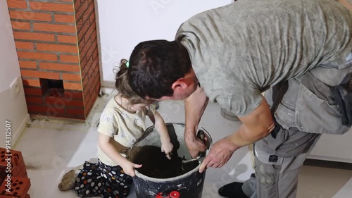 One young handsome man, a builder of the Caucasian race, together with his little daughter, mixes cement mortar in a bucket with the help of spatulas, standing in the evening in a room by the light of
