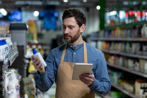 Supermarket employee holding product bottle and tablet while checking inventory. Man in apron working in retail store aisle examining goods for stock control and management. photo